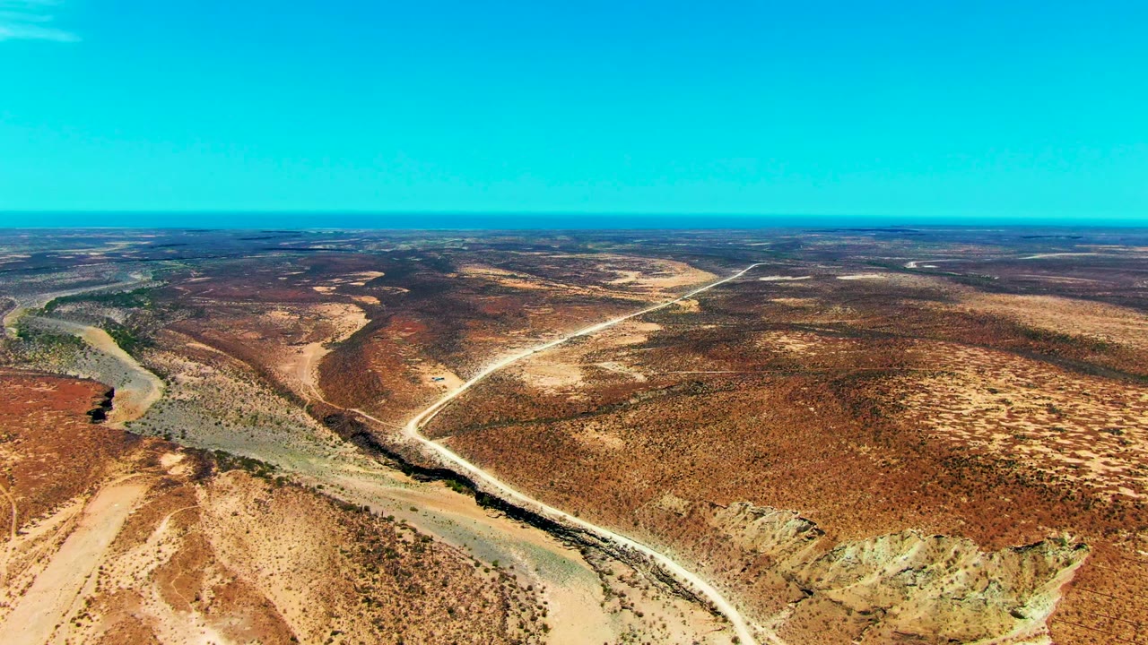 Aerial frame over San Juanico, Baja California Sur showing a pale dirt road beside a dry wash, red-brown scrubland, clear blue sky, and no visible people.