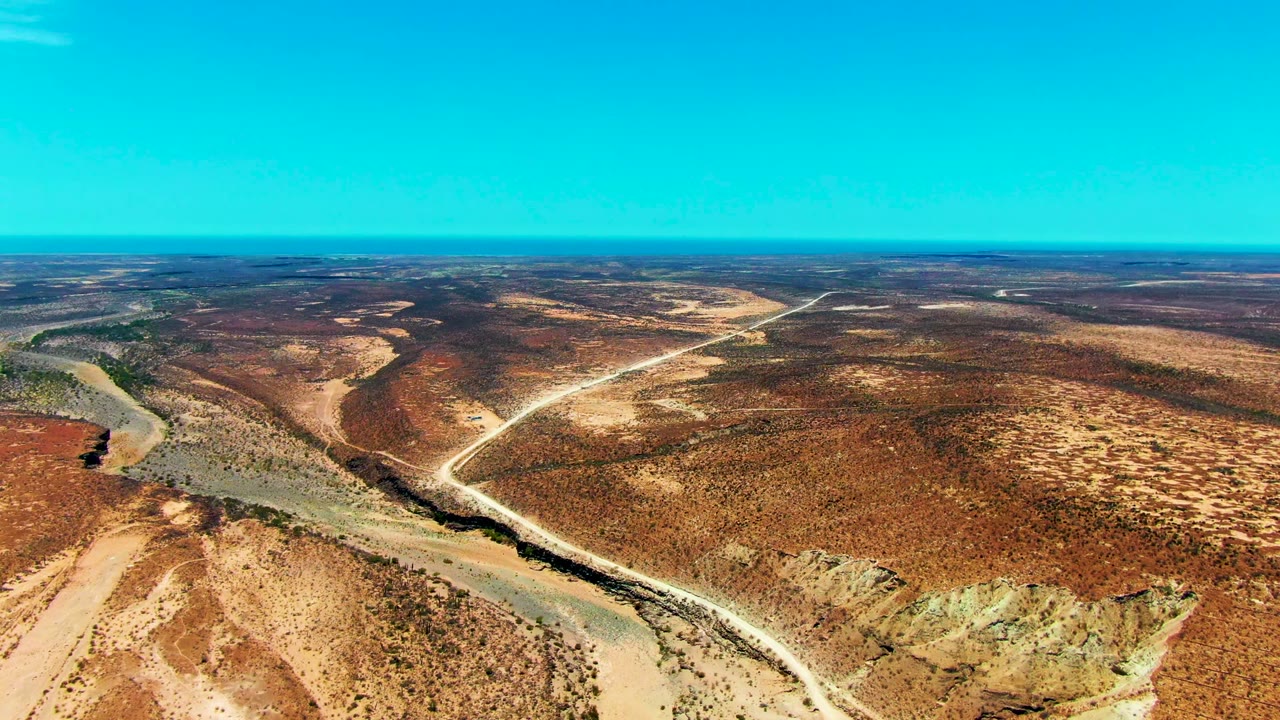 Mid-clip aerial view in San Juanico, Baja California Sur shows a pale road crossing red desert terrain, a dry channel at left, open horizon, and no visible people.
