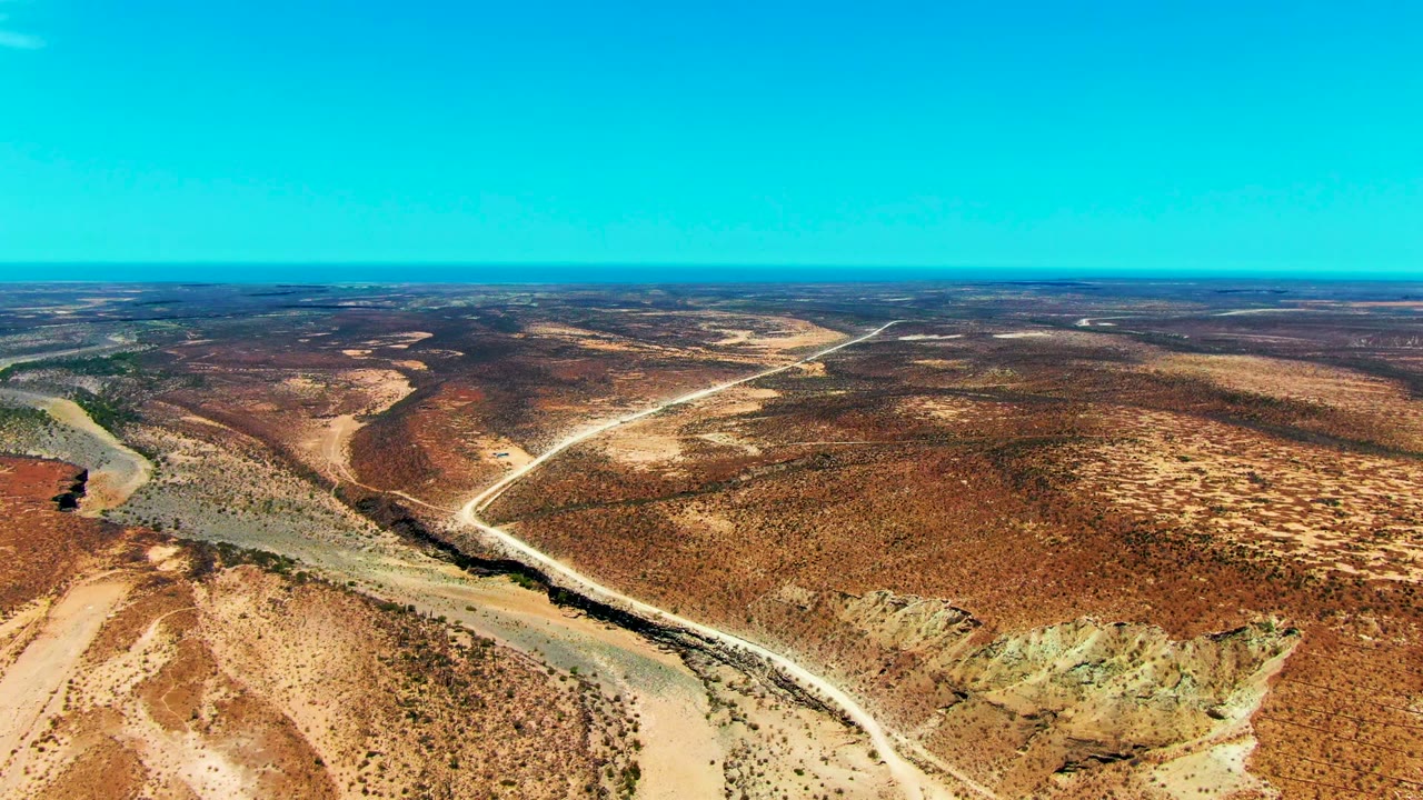 Aerial view over San Juanico, Baja California Sur, with a sandy riverbed crossing the foreground, pale dirt roads through brown scrub, a flat horizon, and no visible people.