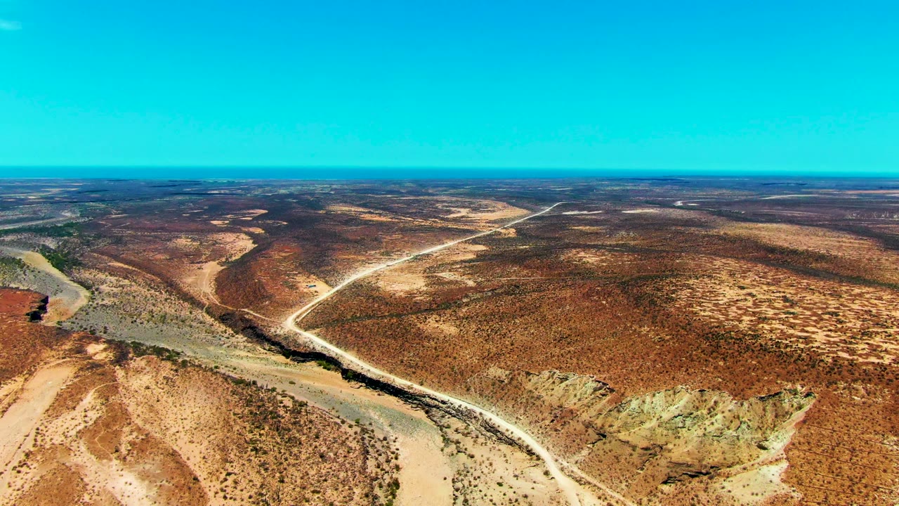 Wide drone view of brown desert scrub with a winding riverbed and crossing dirt track.