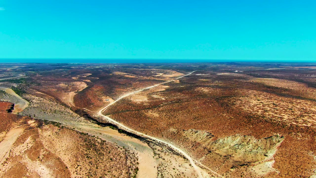 Aerial view over San Juanico, Baja California Sur, with a pale road angling across dry brown scrub, a narrow riverbed along the left side, distant open land, and no visible people.