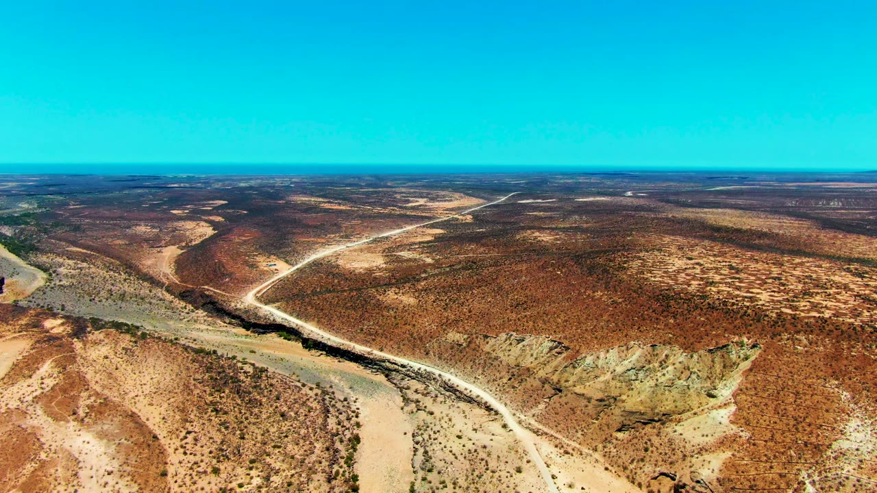 Aerial frame near San Juanico with a pale road running diagonally beside a dry riverbed, scrub patches across the foreground, and no visible people.