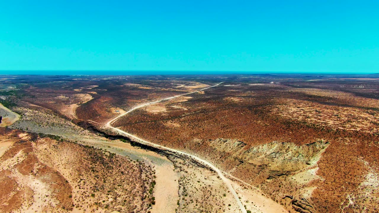 Opening aerial frame over San Juanico, Baja California Sur, showing a pale dry riverbed curving through reddish desert scrub with a dirt road crossing the terrain and no visible people.