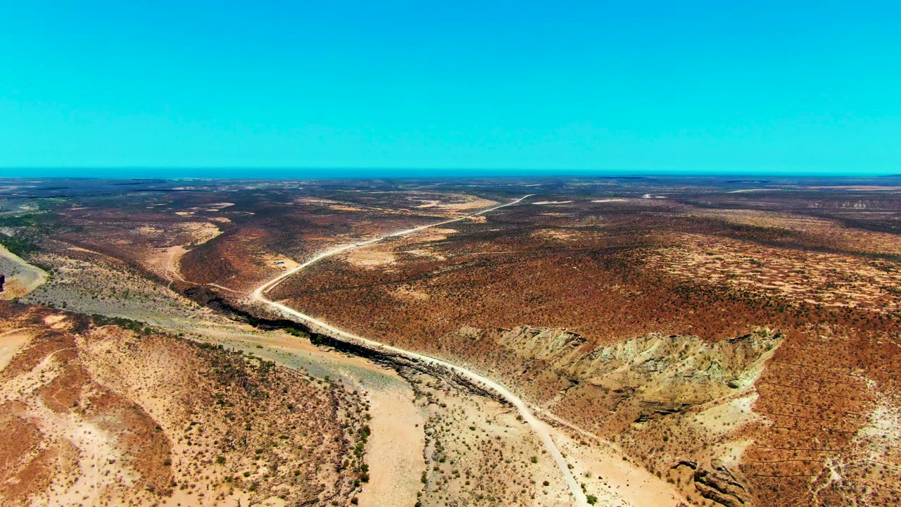 Wide aerial over arid terrain with pale riverbed, dirt track, and low scrub.