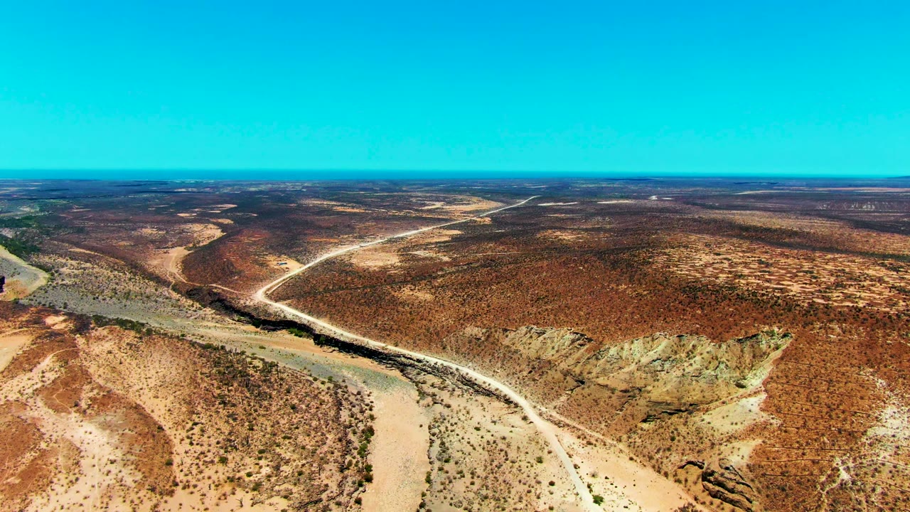 High oblique view in San Juanico, Baja California Sur, with a winding dry wash at lower left, a light dirt road crossing the middle distance, reddish-brown scrubland, and no visible people.