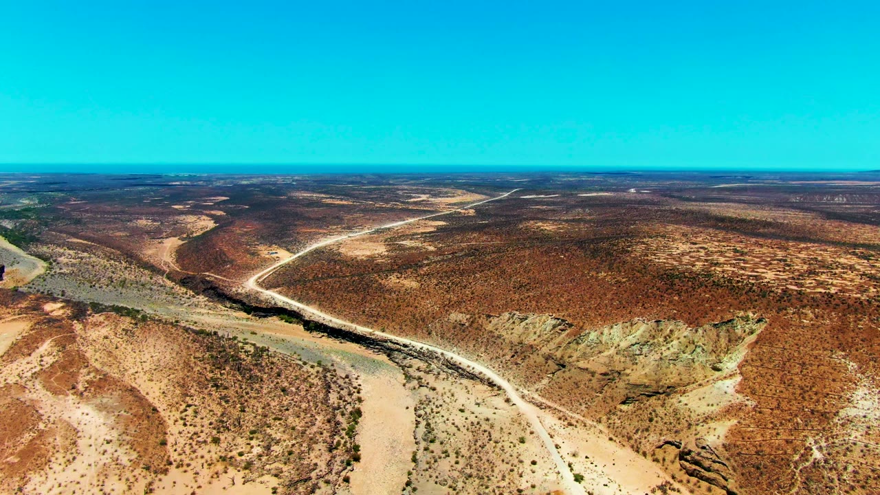 Drone-like desert panorama with dry river channel and pale road through scrub.