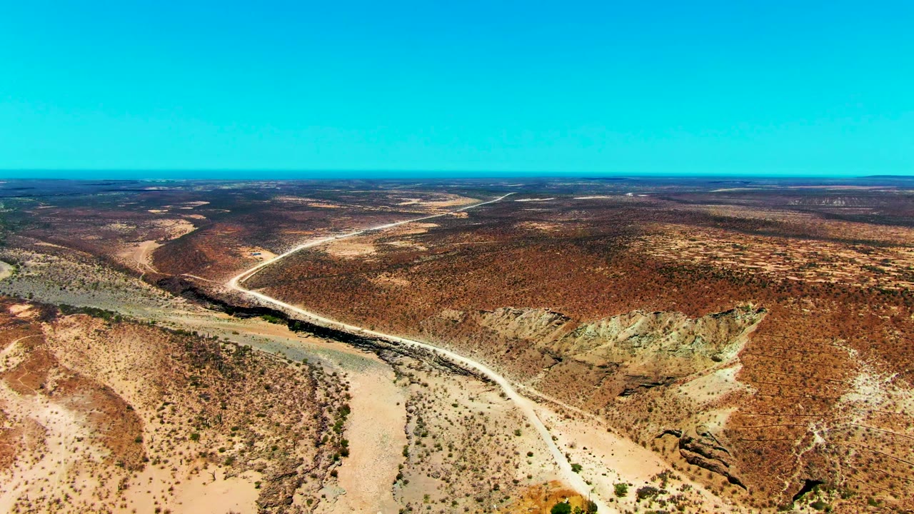 Opening aerial frame over San Juanico, Baja California Sur, with a pale dirt road curving beside a dry wash across brown scrubland and no visible people.