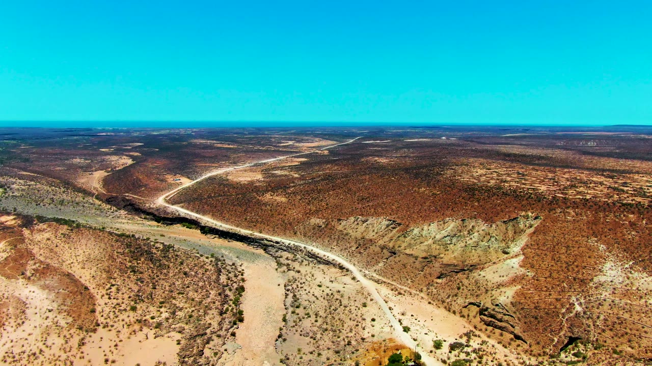 Second aerial frame in San Juanico, Baja California Sur, showing the road continuing through reddish-brown desert and crossing near a dry channel with no visible people.