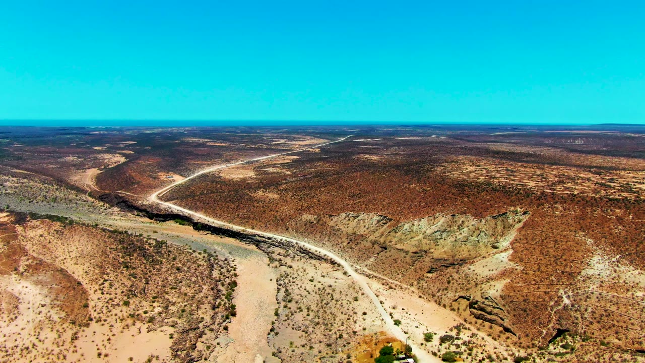 Mid-clip aerial frame over San Juanico, Baja California Sur, with the pale road bending from the lower landscape toward the distant horizon and no visible people.