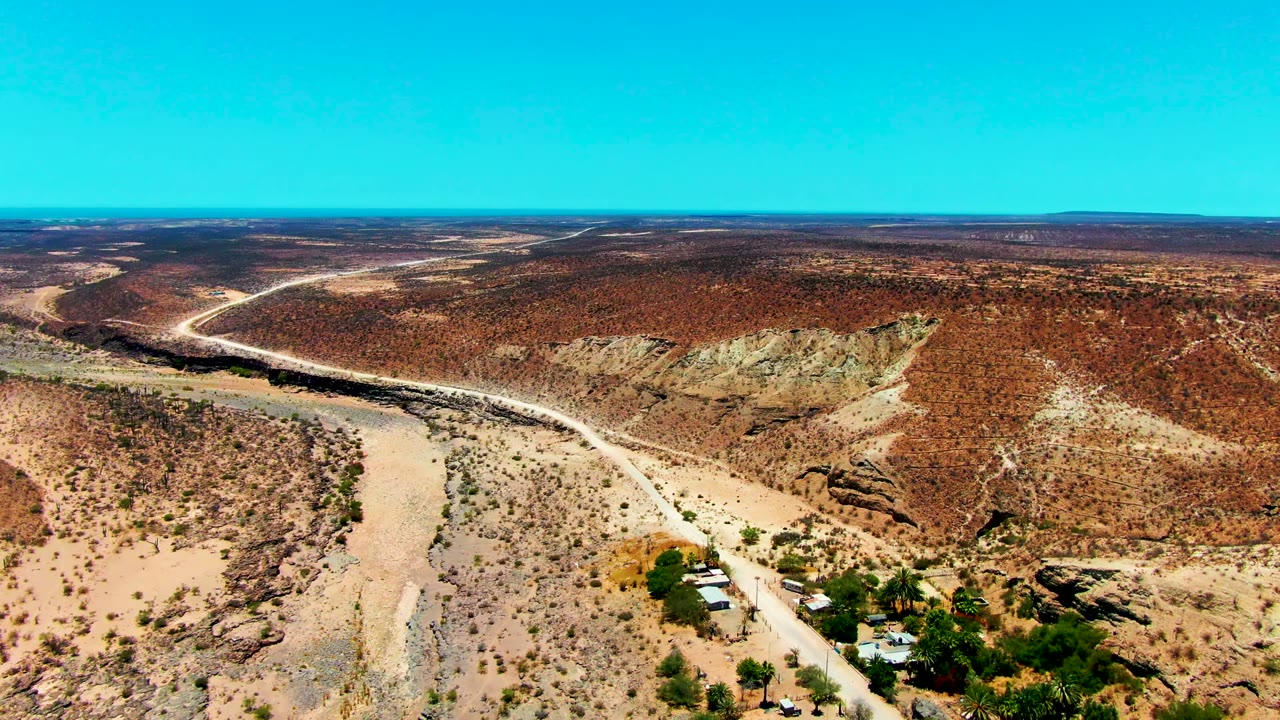 Aerial frame from San Juanico, Baja California Sur showing a pale dirt road curving beside a dry riverbed, scattered shrubs, low rust-colored hills, small structures near the lower edge, and no visible people.