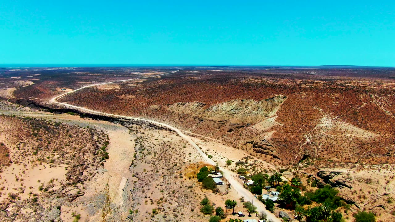 Aerial frame from San Juanico, Baja California Sur with the curving dirt road more central, the dry riverbed running through rocky ground, sparse vegetation across the slopes, and no visible people.