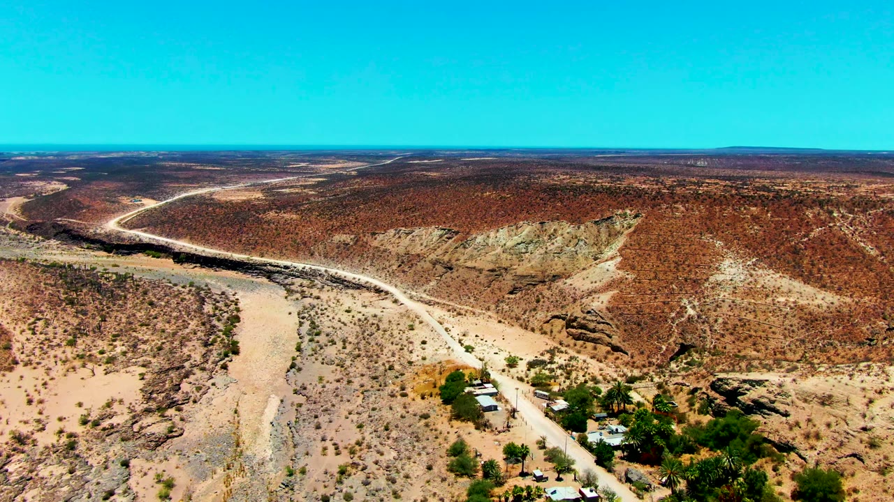 Aerial frame from San Juanico, Baja California Sur showing the road and dry river channel passing below scrub-covered hills, with a small cluster of buildings and trees near the lower frame and no visible people.