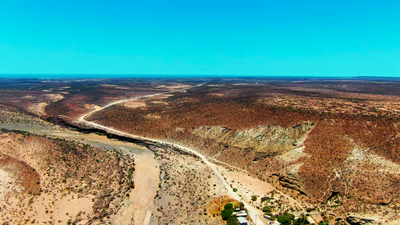 Aerial frame from San Juanico, Baja California Sur showing a pale dirt road curving through reddish arid hills with sparse scrub, a small settlement at the lower edge, and no visible people.