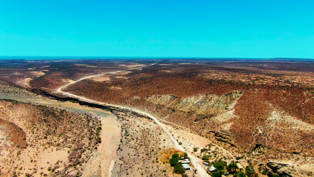 San Juanico aerial frame showing a pale dirt road beside a dry wash, reddish-brown slopes, scattered desert shrubs, small buildings near the bottom edge, and no visible people.