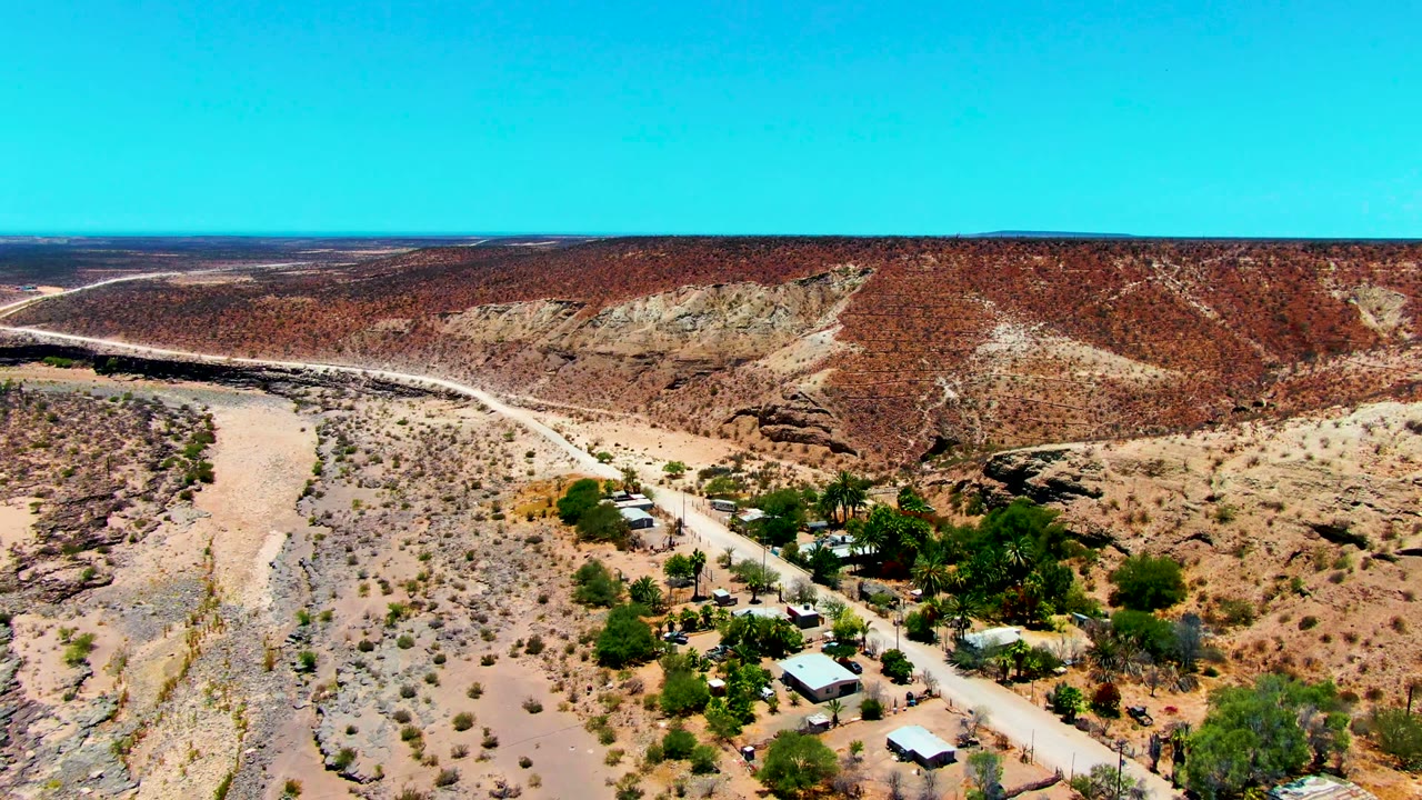 Aerial frame over San Juanico, Baja California Sur, showing a broad dry riverbed beside a pale dirt road, small buildings, green trees, red hills, and no visible people.