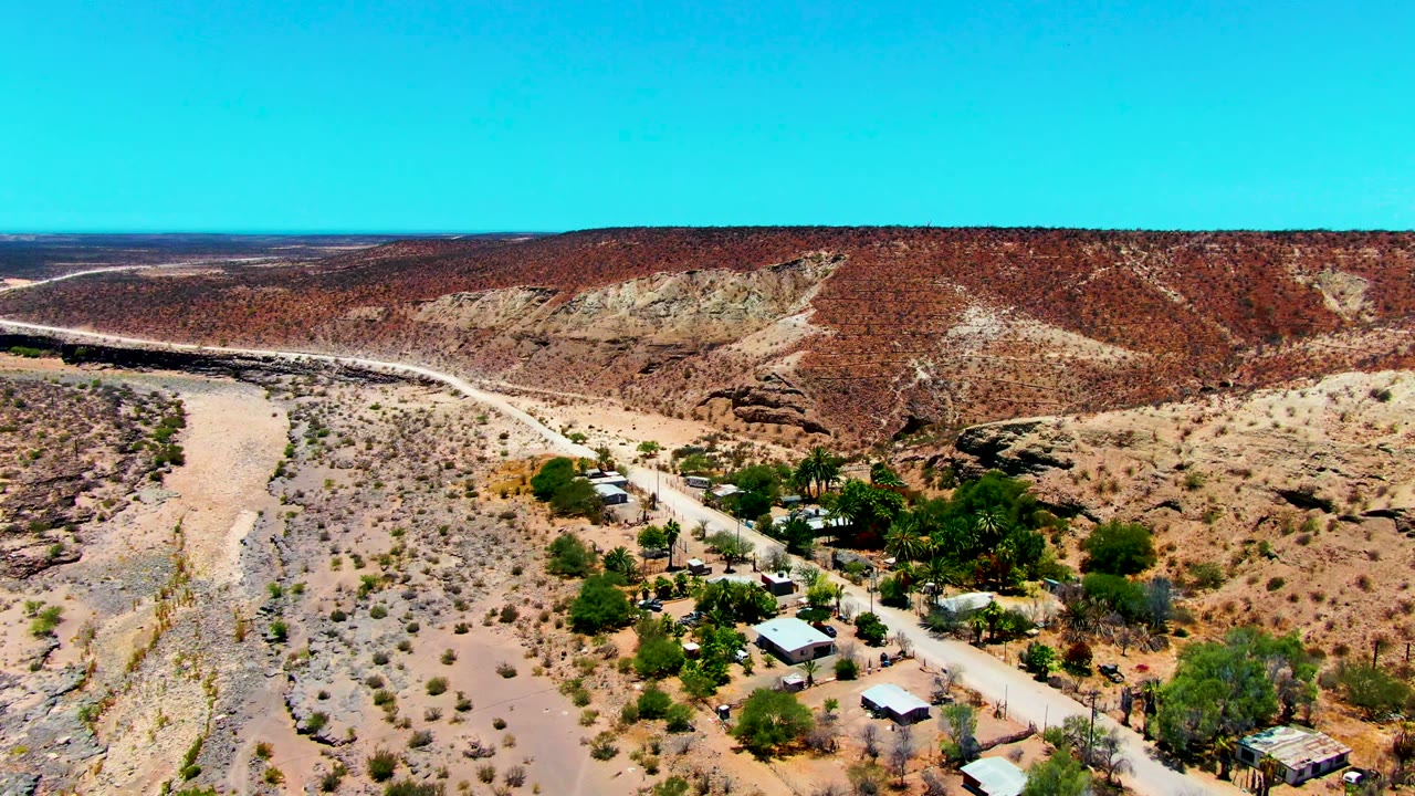 Aerial frame from San Juanico, Baja California Sur, showing the dry river channel and road curving past scattered buildings and trees beneath a clear sky, with no visible people.