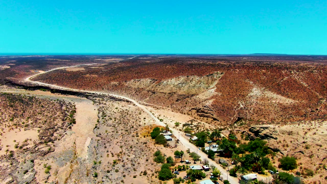 Opening aerial frame over San Juanico, Baja California Sur, with a pale road curving through dry terrain, light-roofed buildings near scrub vegetation, red-brown hills, blue sky, and no visible people.