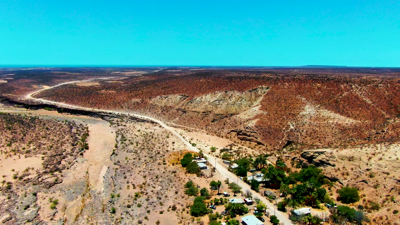 High aerial view of desert scrub, a curving road, rocky slopes, and scattered buildings.