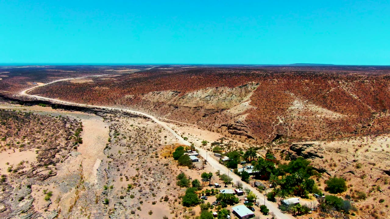 San Juanico aerial frame with a pale road tracing the base of red and tan hills, scattered scrub across sandy ground, a compact group of buildings near the lower edge, blue sky, and no visible people.