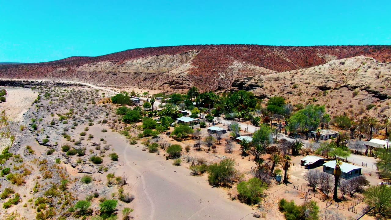 Aerial frame over San Juanico, Baja California Sur, showing sandy desert ground, scattered shrubs, small buildings, palms, rocky hills, blue sky, and no visible people.