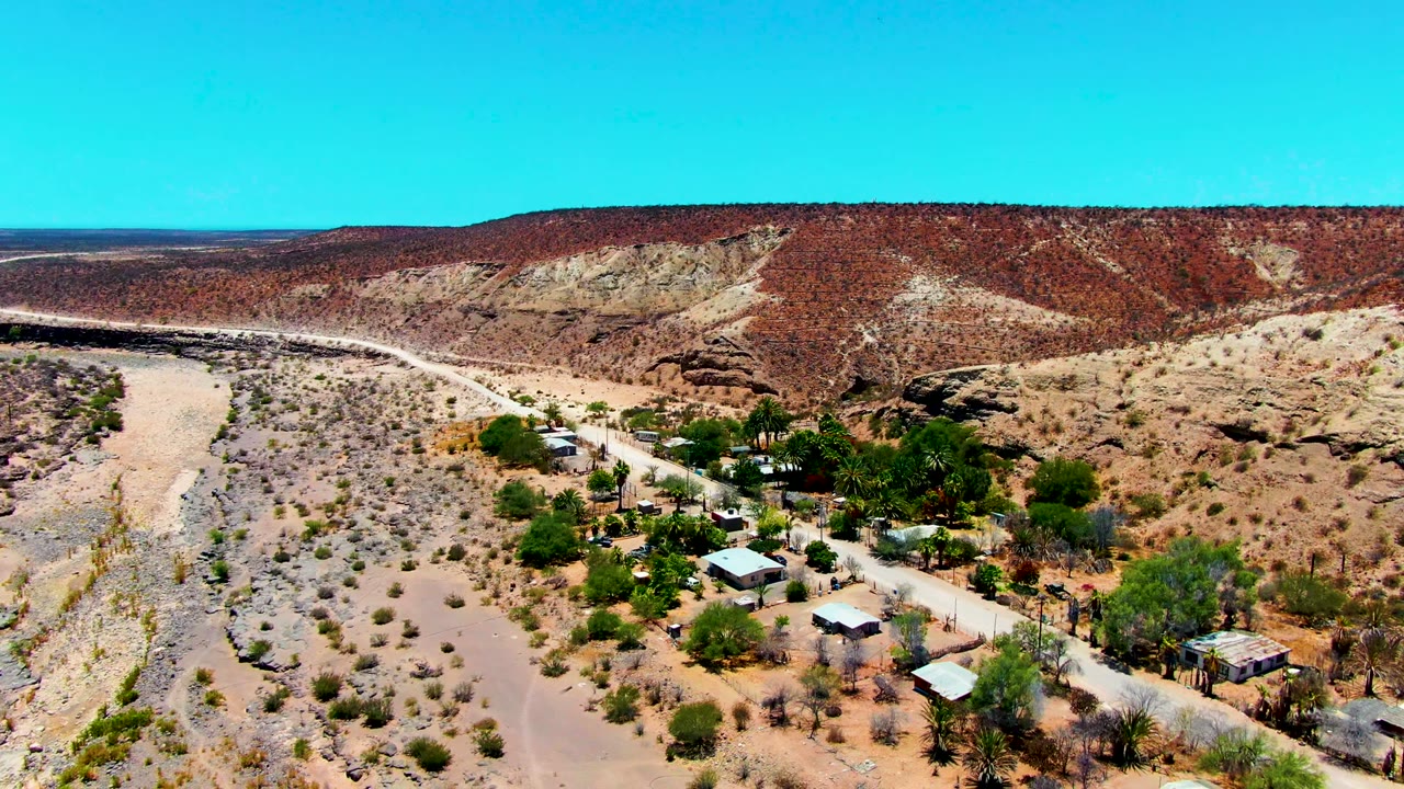 Aerial frame over San Juanico, Baja California Sur showing a sandy wash at left, small buildings and palms near a road, red rock hills beyond, and no visible people.
