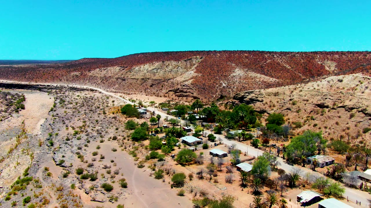Drone frame over San Juanico showing clustered palms and low structures beside a dusty road, with open desert wash and red hillside in the background, no visible people.