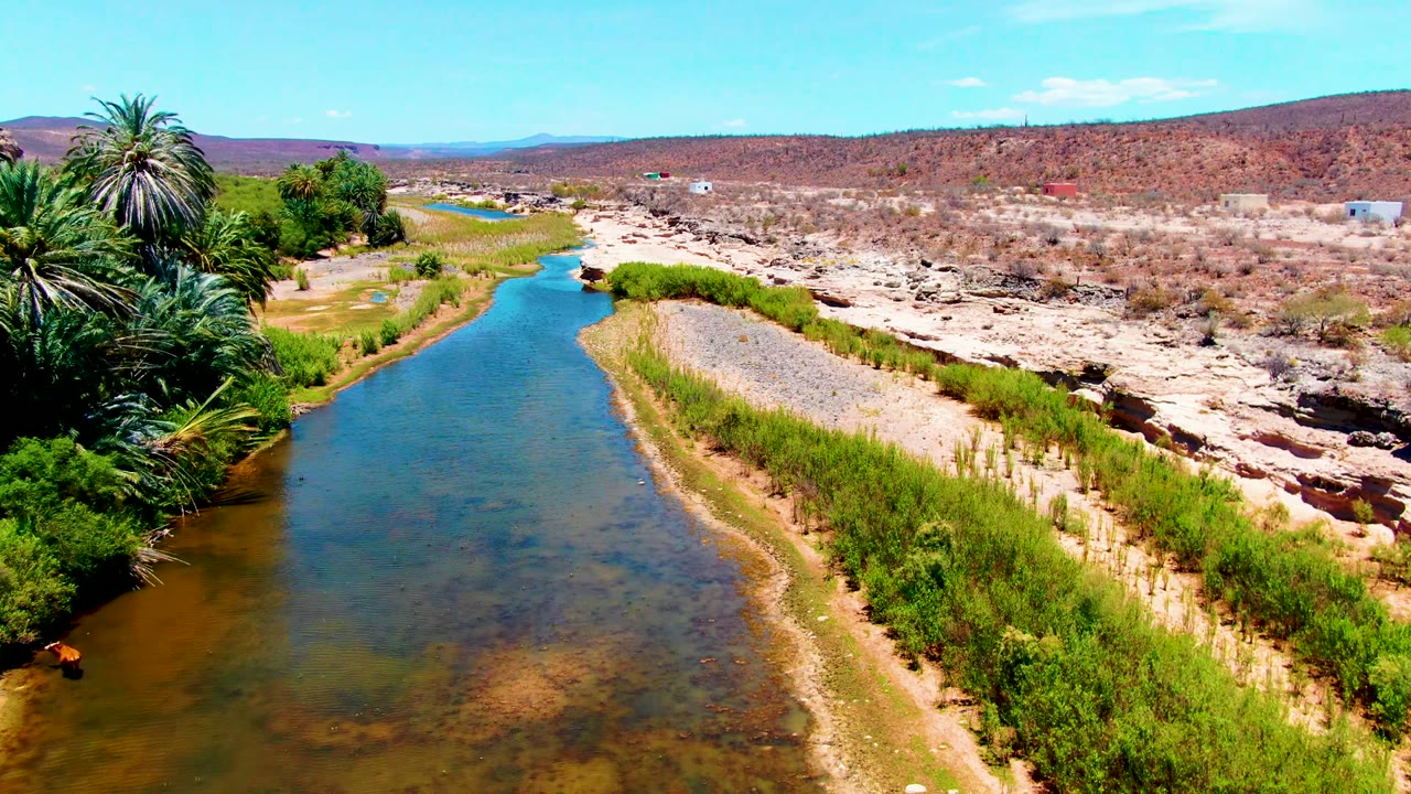 Aerial view in San Juanico, Baja California Sur, showing a blue-green river curving past palms, gravel banks, dry rocky ground, and no visible people.