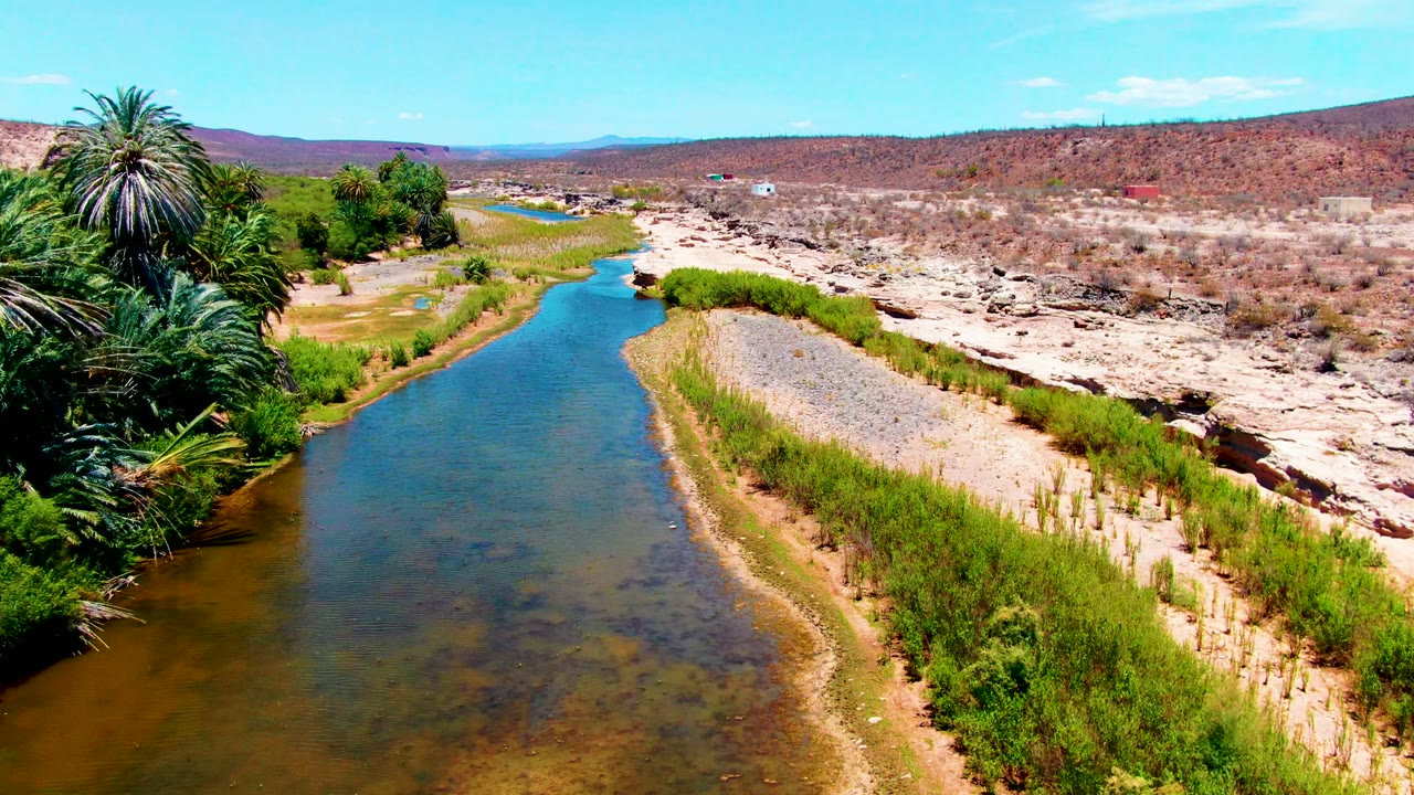 River channel with green banks, palms, gravel, and desert hills.