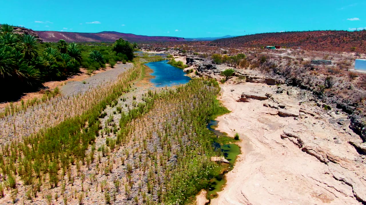 Aerial river channel with green planting rows, palms, rocks, hills, and blue sky.