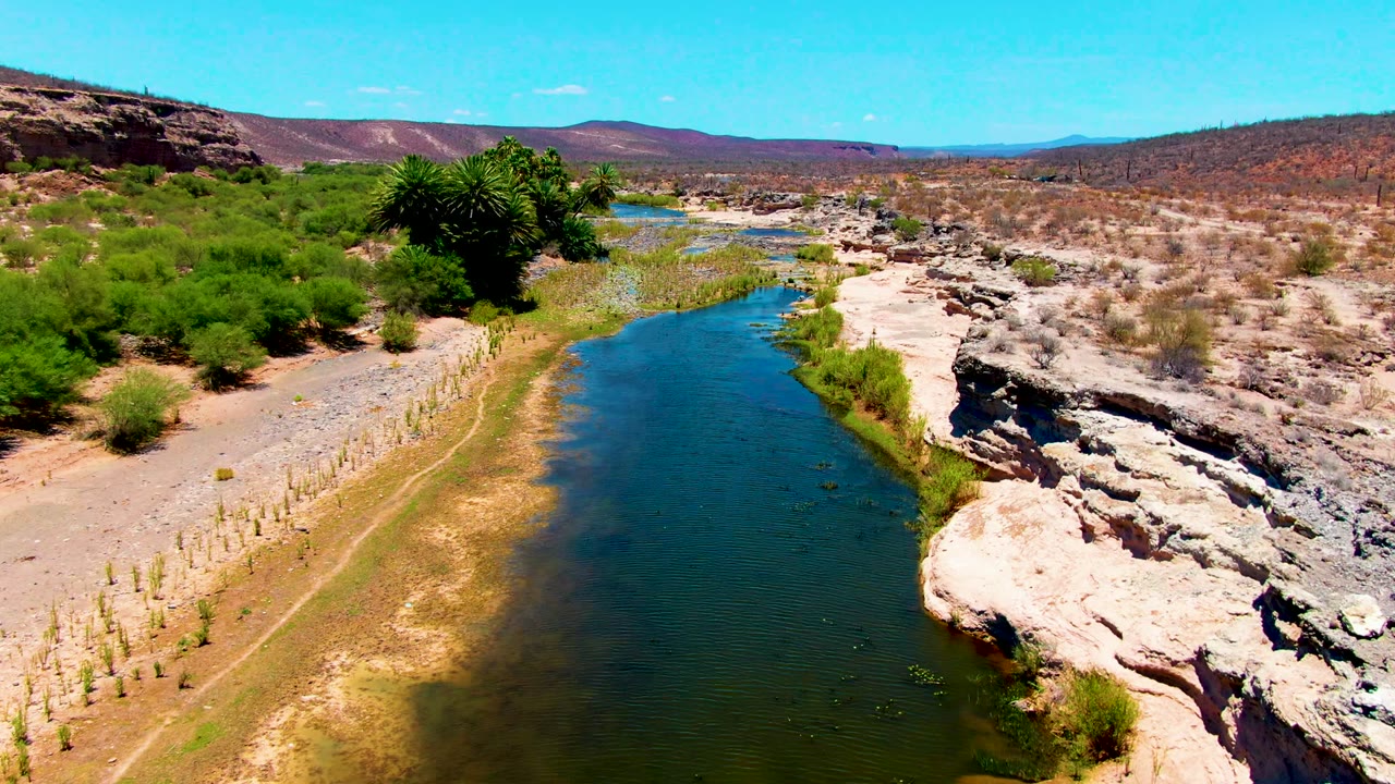 Aerial view in San Juanico, Baja California Sur, Mexico, showing a blue-green river bordered by pale rock shelves, scrubby banks, distant hills, and no visible people.