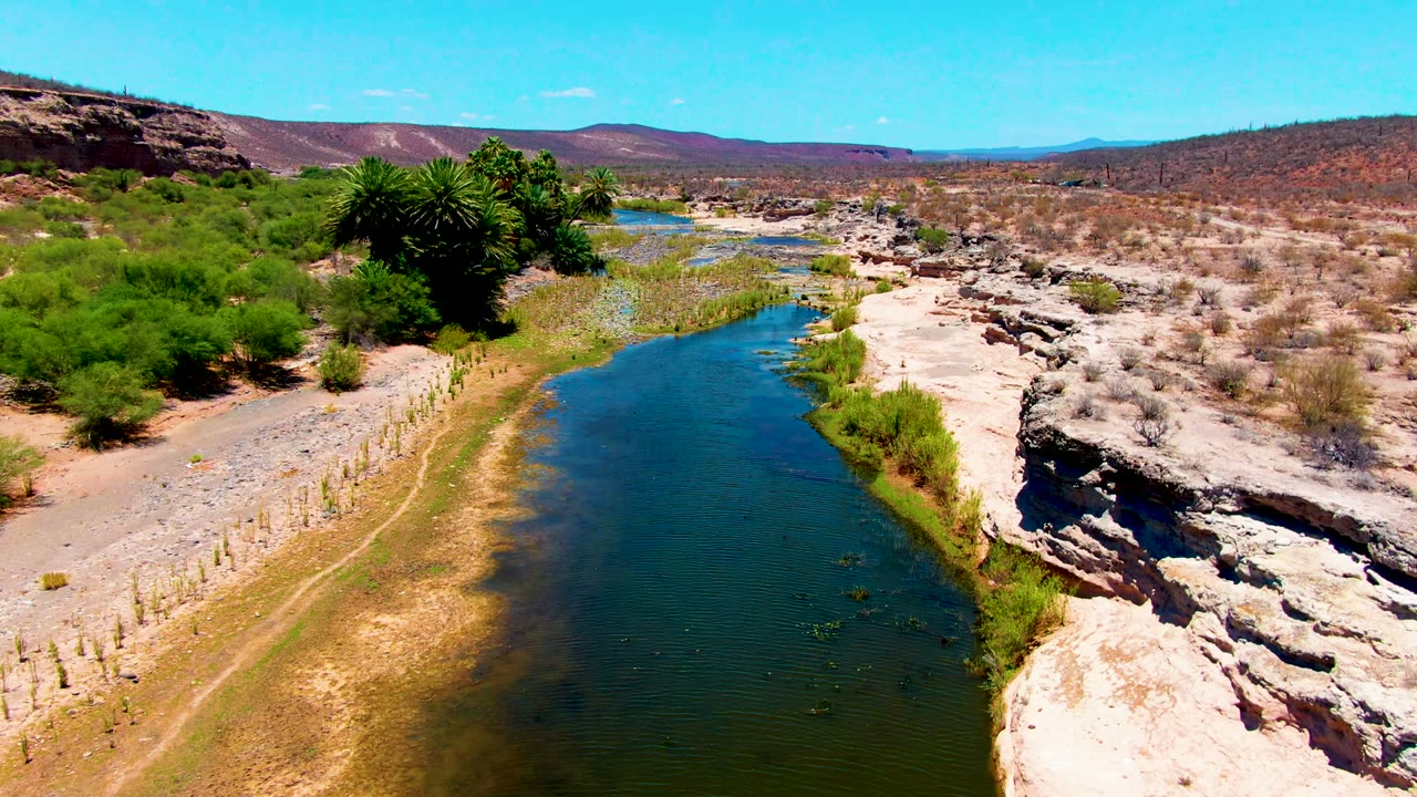 Aerial view over San Juanico terrain with blue water in the lower frame, rough pale rocks along the right bank, clustered green shrubs on the left, and no visible people.