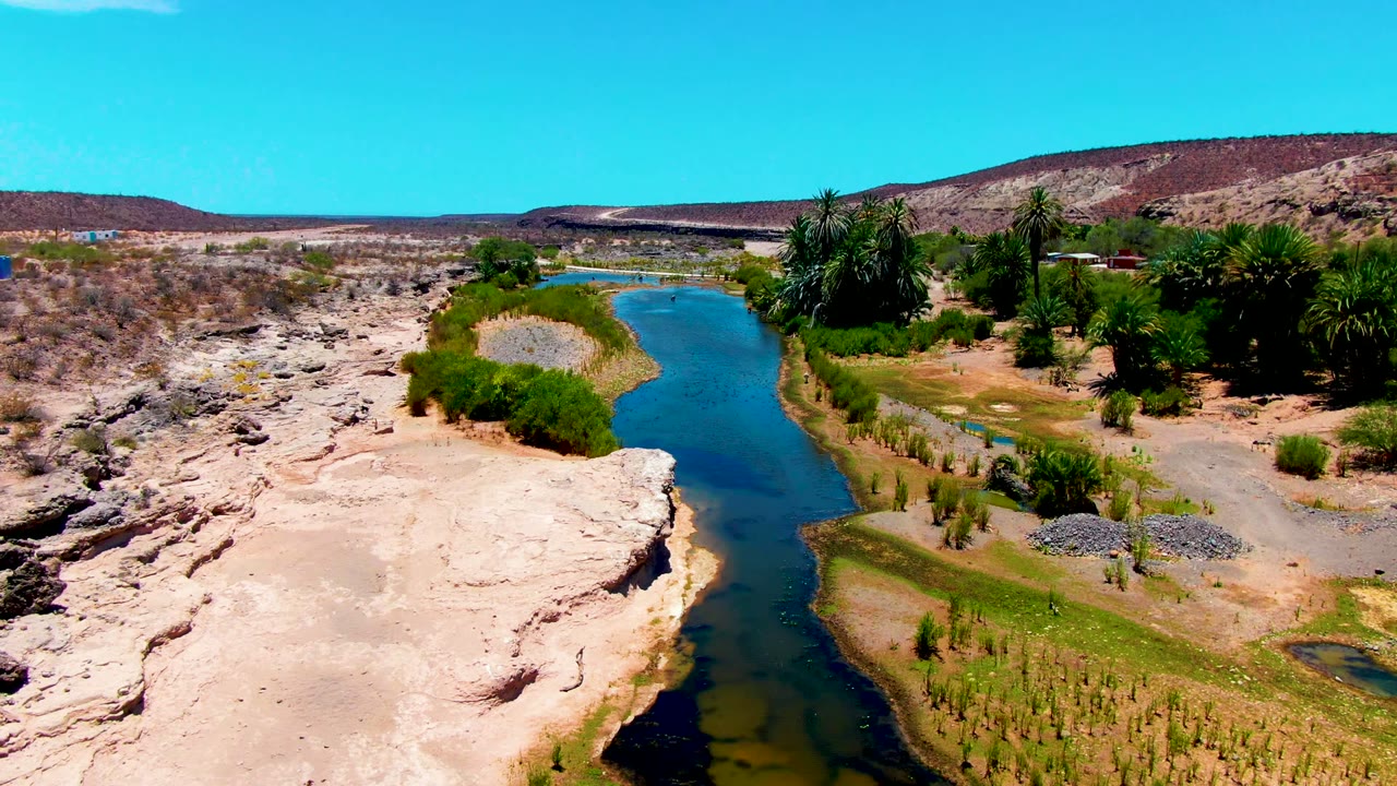 Aerial frame over San Juanico, Baja California Sur showing a narrow blue-green water channel between pale rock, scrub, palms, low hills, and clear sky, with no visible people.