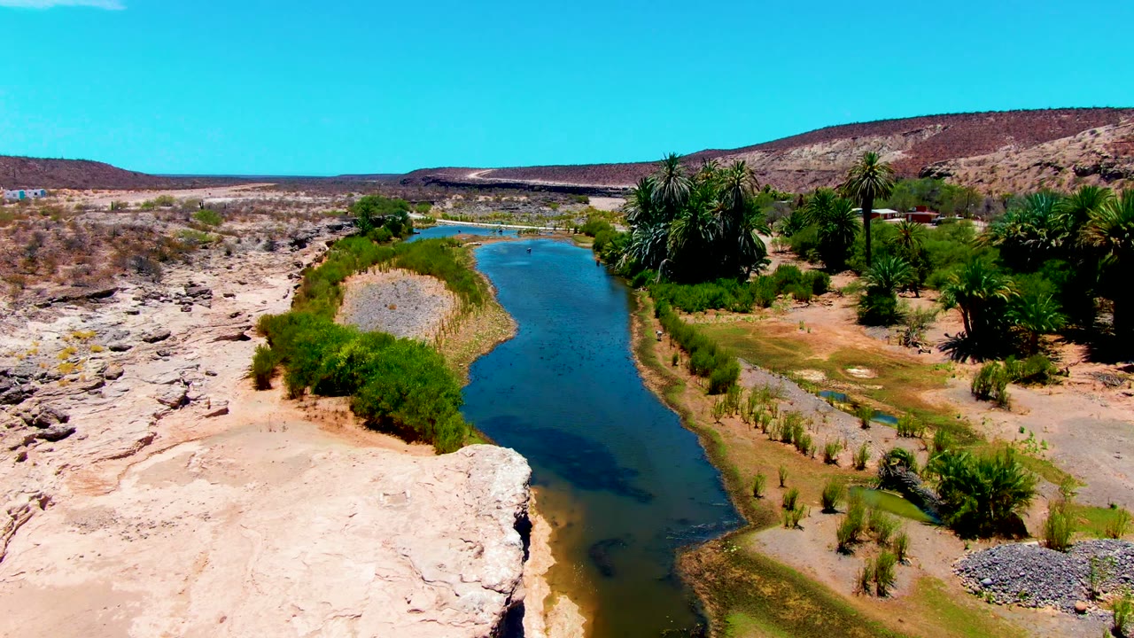 San Juanico, Baja California Sur appears from above with a curving blue channel, green banks, palms to the right, pale rocky desert to the left, and no visible people.