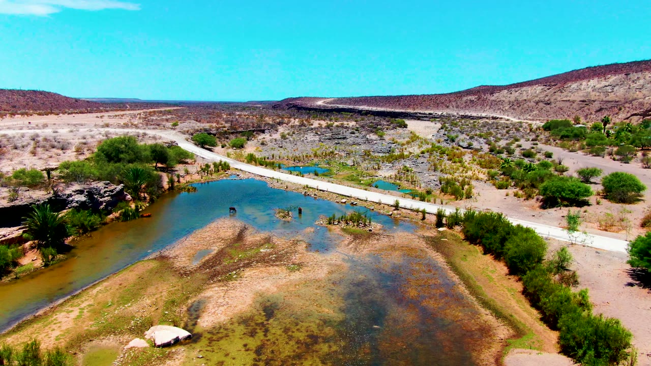 Drone view of river water, sandbar, trees, and a road across dry terrain.