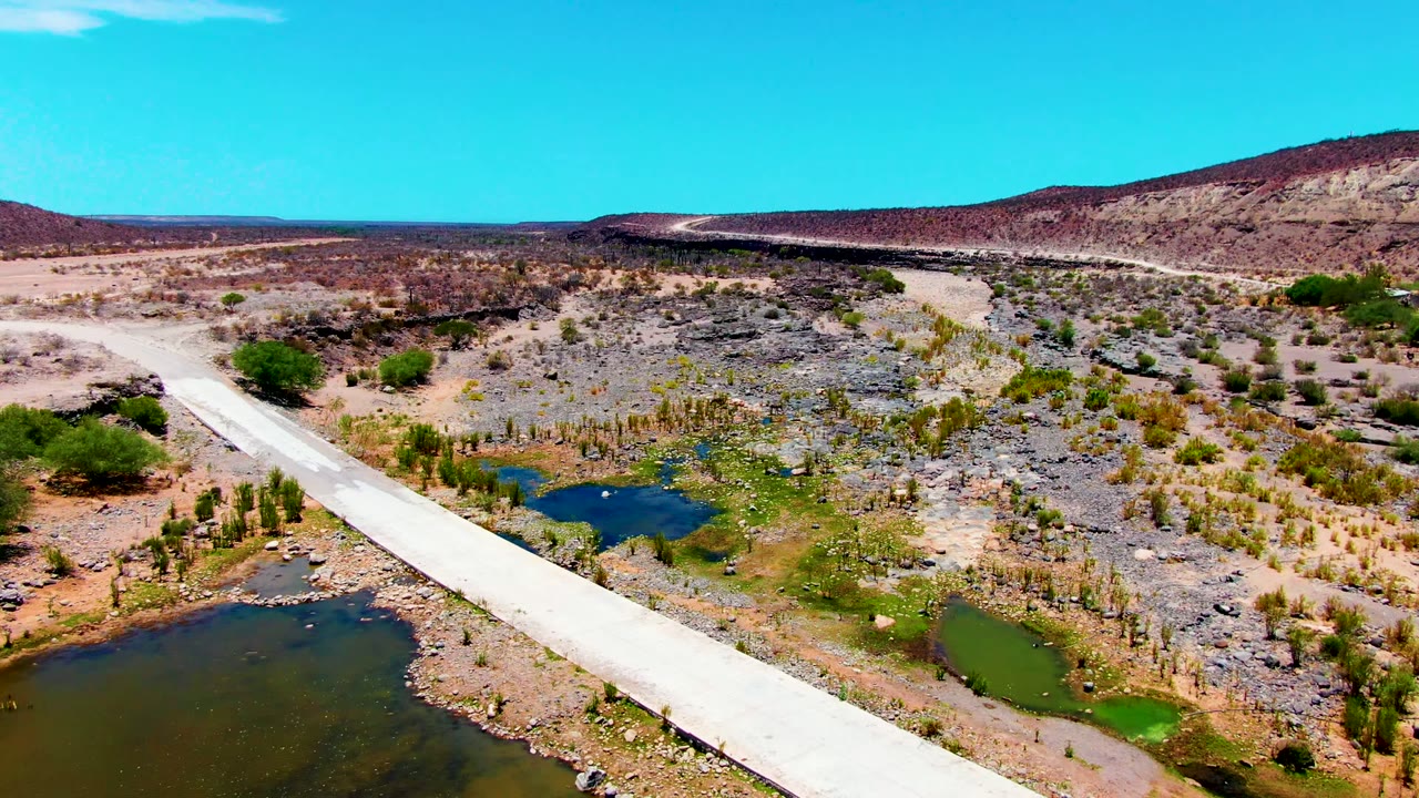 Aerial frame over San Juanico, Baja California Sur showing a pale bridge entering from the lower left across small water pools, rocky desert ground, low hills, clear blue sky, and no visible people.