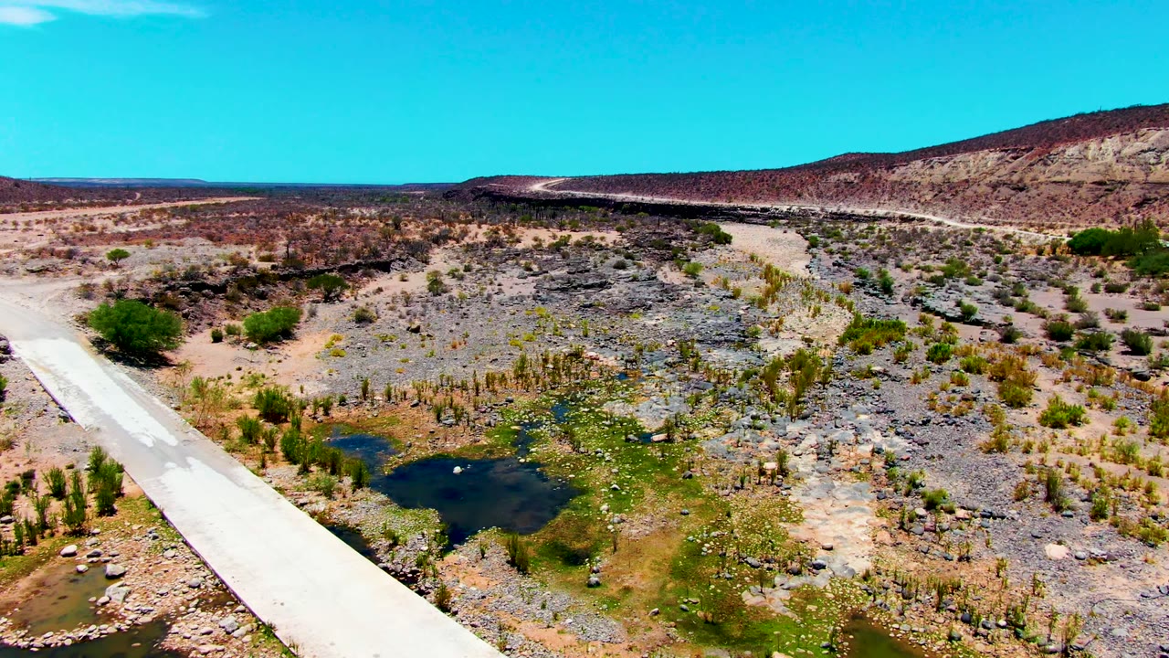 Aerial frame over San Juanico, Baja California Sur where the bridge shifts toward the left edge, revealing more rocky wash, small water pools, sparse vegetation, low hills, clear blue sky, and no visible people.