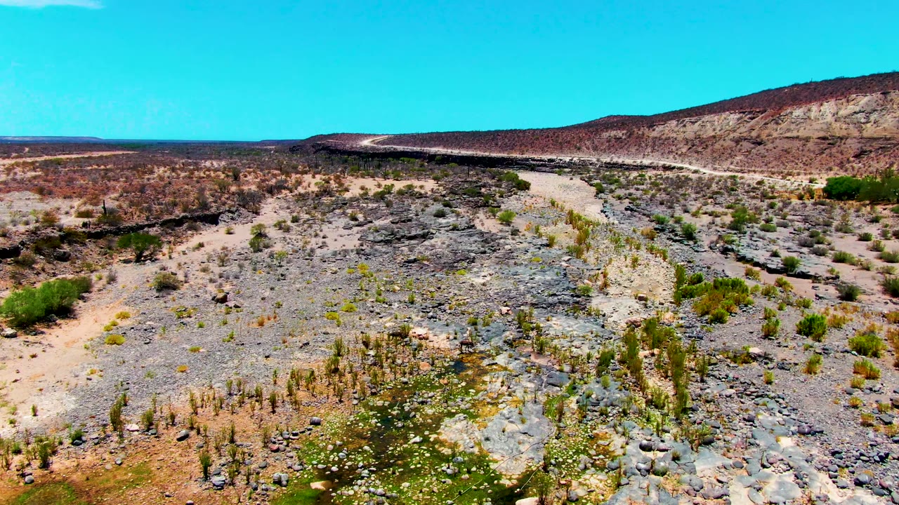 Aerial view of rocky desert terrain near San Juanico, Baja California Sur, with pale wash channels, scattered shrubs, a low reddish hillside, and no visible people.