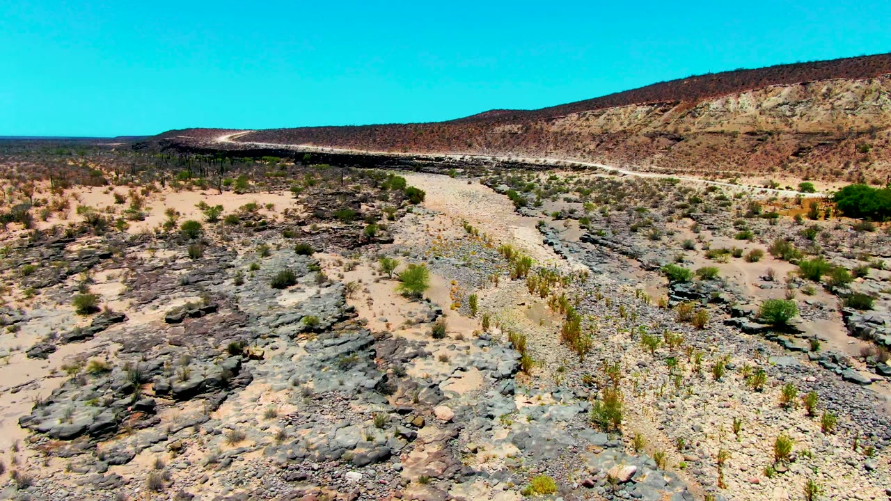 Opening aerial view in San Juanico, Baja California Sur, showing a pale rocky path between dark stone shelves, scattered desert plants, a distant ridge, and no visible people.