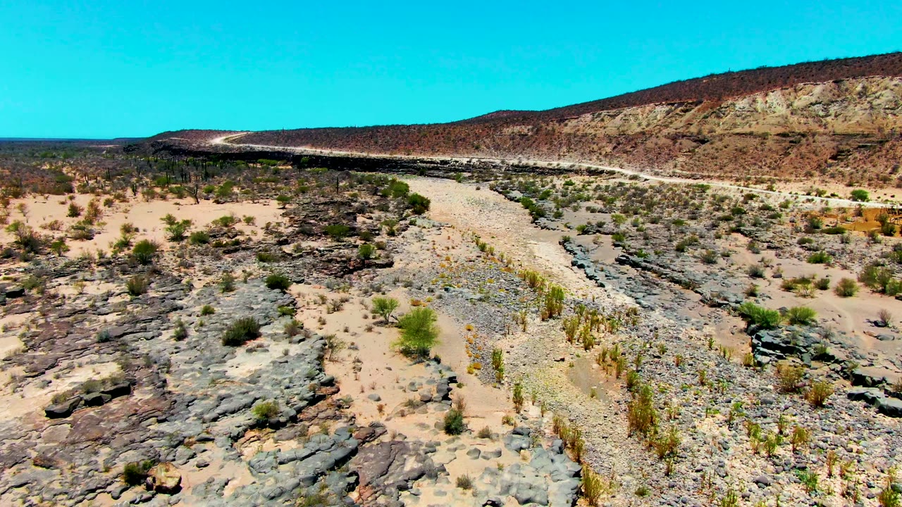 Mid-clip aerial frame in San Juanico, Baja California Sur, with the pale path running diagonally through rocky desert ground, scrub patches, cactus-like plants, a ridge line, and no visible people.