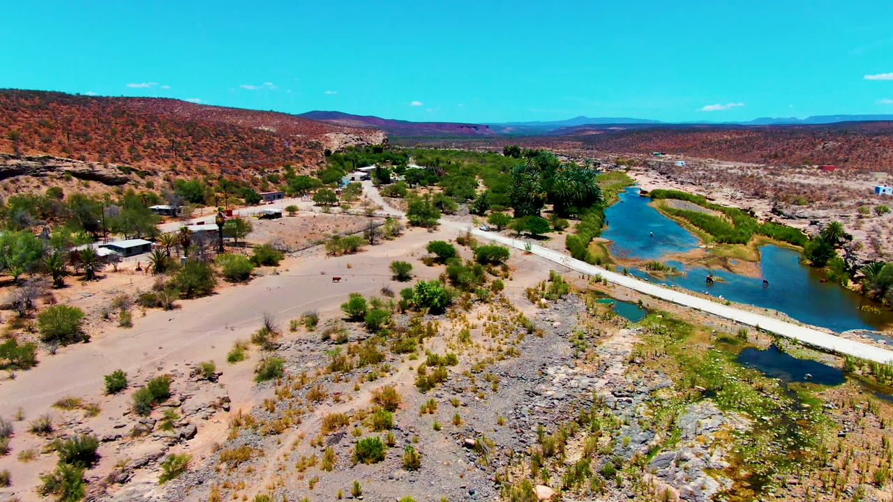 Aerial view over San Juanico, Baja California Sur, showing a blue desert river, pale bridge, rocky wash, scattered vegetation, small structures, red hills, and no visible people.