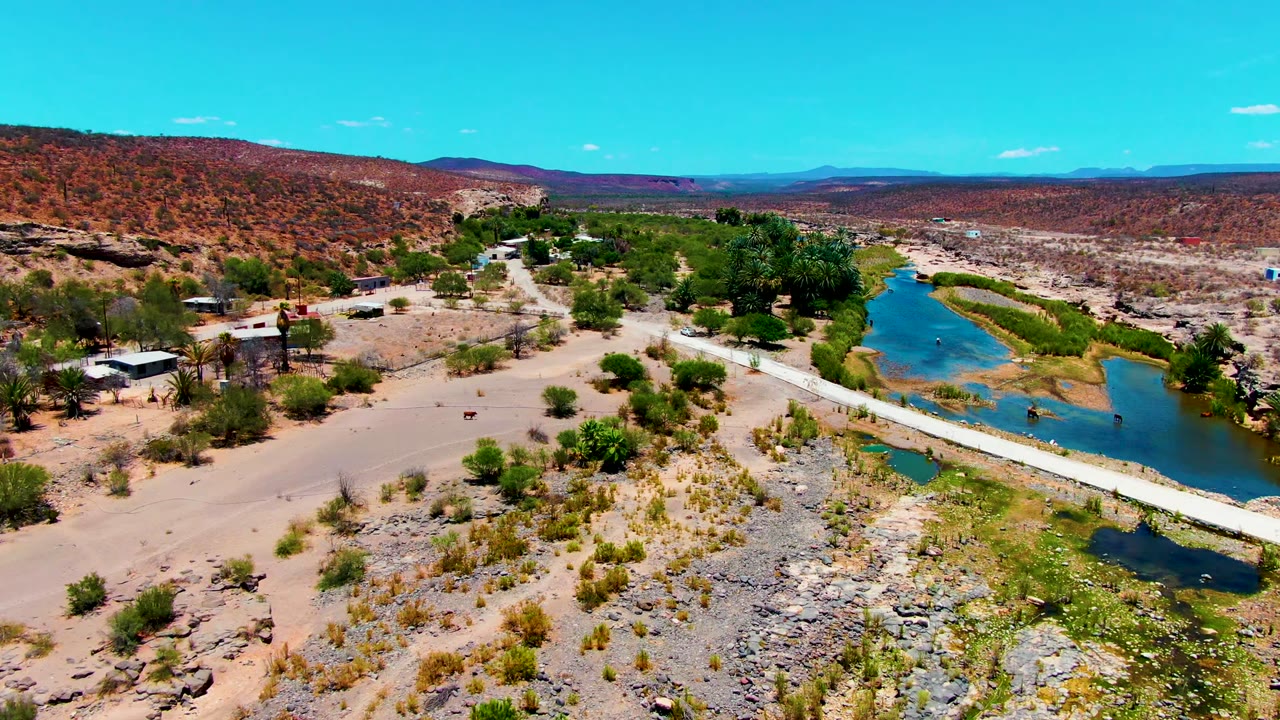 San Juanico aerial frame showing the river curving past a small settlement, the bridge spanning the water, dry rocky foreground, reddish hills beyond, and no visible people.