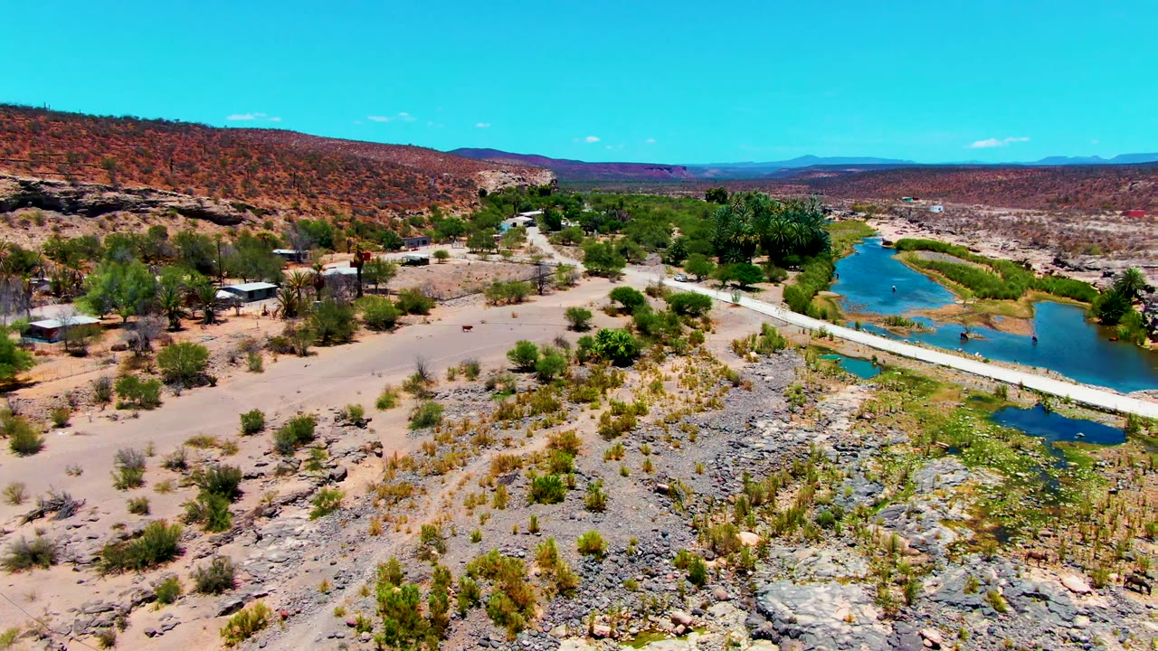 Aerial frame over San Juanico, Baja California Sur, showing a pale road crossing beside a small blue water body, rocky desert foreground, scattered buildings, distant hills, and no visible people.