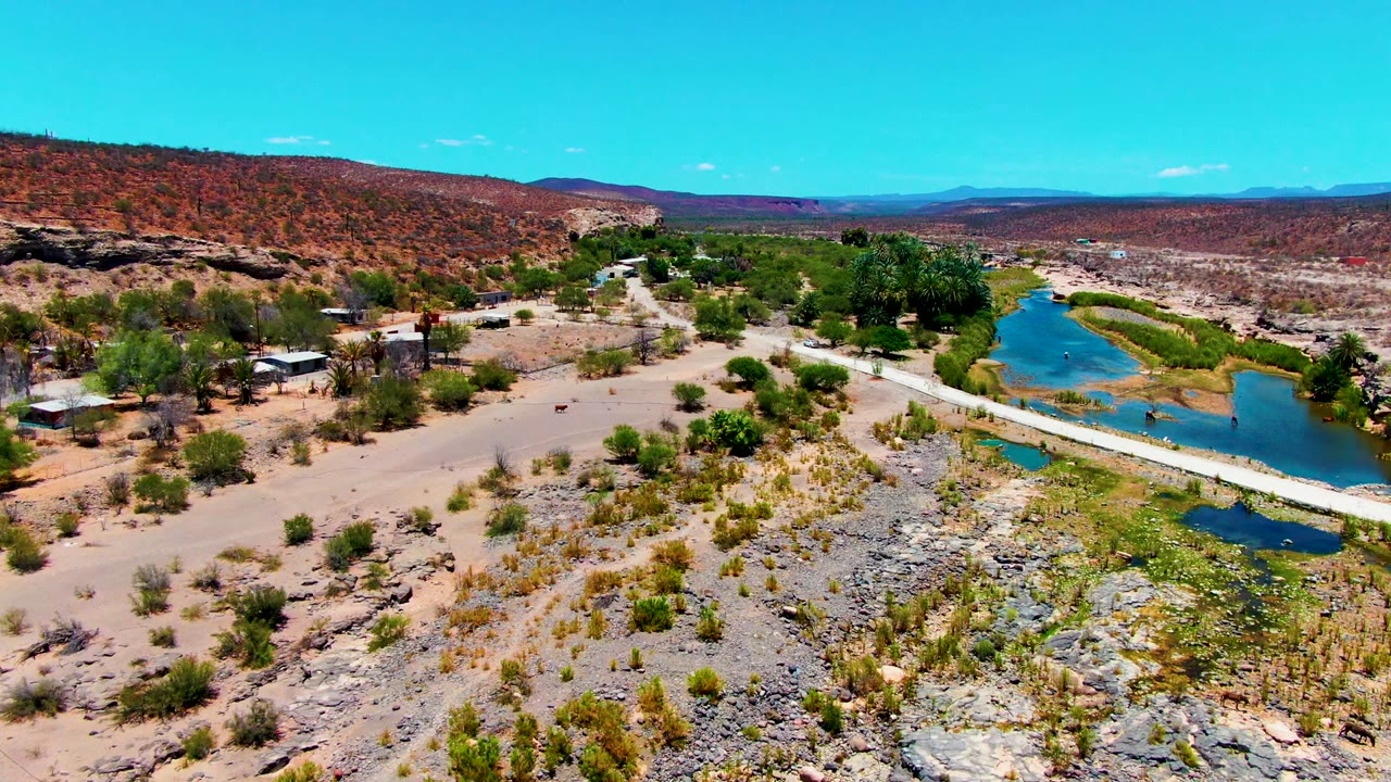 Wide aerial over a desert road, water channel, scrub, and low hills.