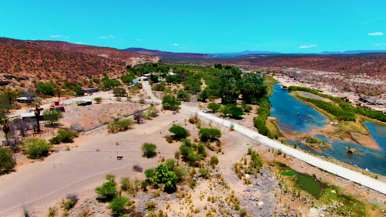 Drone view of a river channel beside sandy ground and scattered buildings.