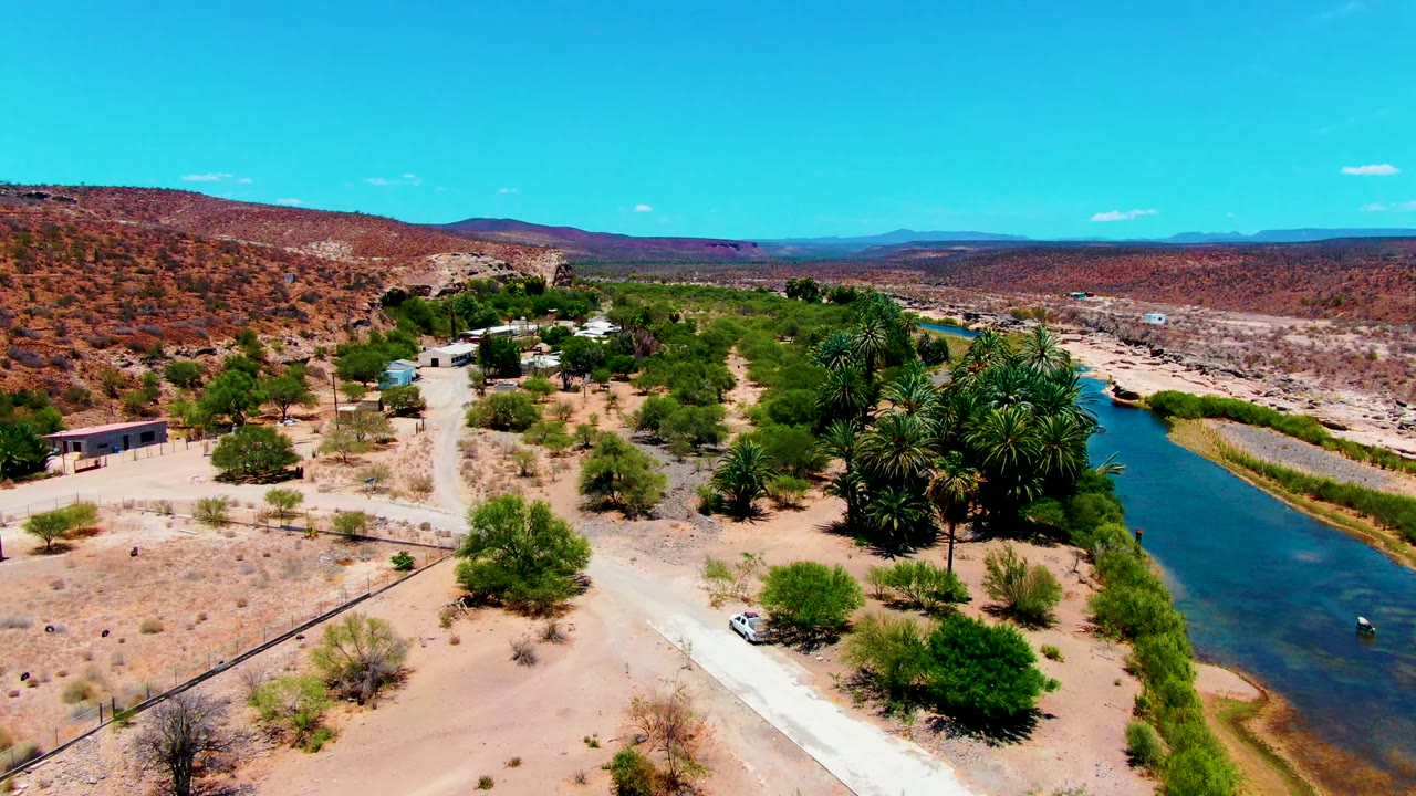 Aerial frame from San Juanico, Baja California Sur showing a sandy road, a blue river channel, palms, scrub, low hills, and no visible people.