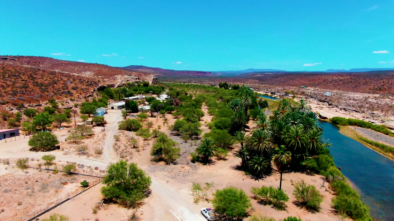 A San Juanico aerial frame showing sandy tracks crossing dry terrain, dense palms near the river, scattered buildings, red-brown hills, and no visible people.