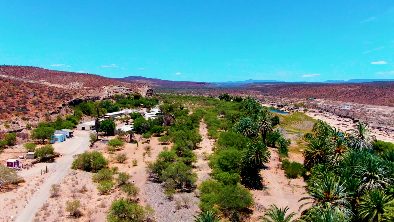 Drone view of sandy desert tracks, palms, scattered buildings, hills, and distant mountains.