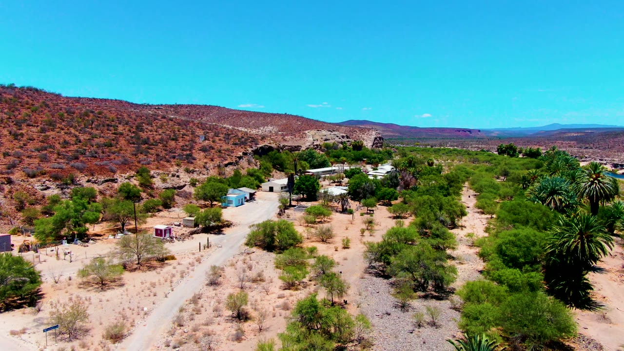 Early aerial frame over San Juanico, Baja California Sur, showing a pale dirt road through sandy terrain, scattered green trees, palms, small buildings, brown hills, distant mountains, and no visible people.