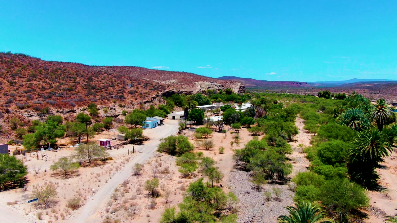 Elevated view of desert scrub, small buildings, a dirt road, palms, and distant mountains.