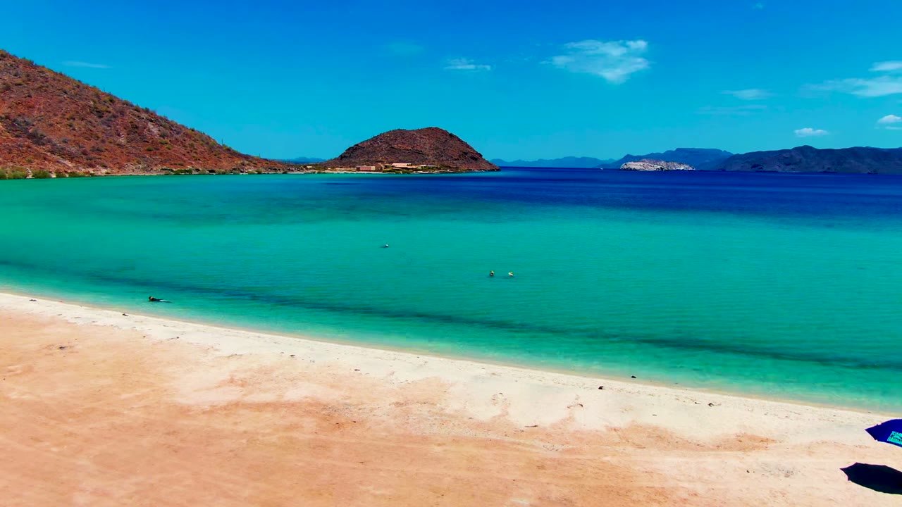 At Playa Santispac, pale sand curves across the foreground while turquoise water, tiny swimmers, brown hills, and a bright blue sky fill the frame.